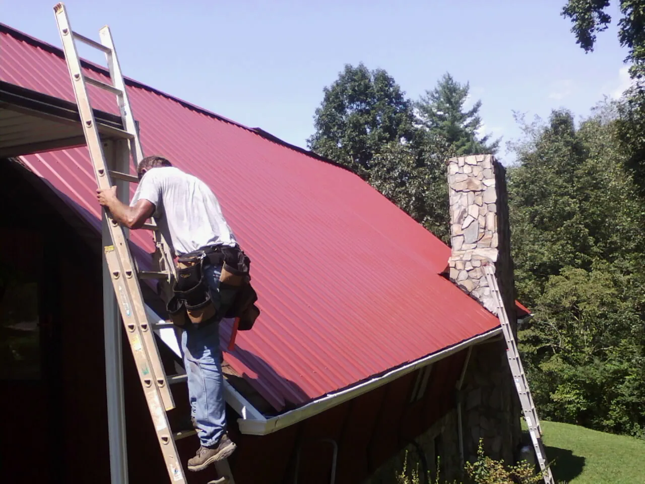 Roofer on a ladder beside a red metal roof and stone chimney — Soffit & Fascia Repair by Roofing Brothers in Chincoteague Island